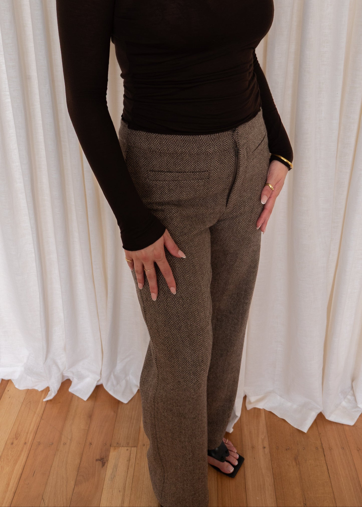Person wearing a brown long-sleeve top and brown pants against a white curtain background