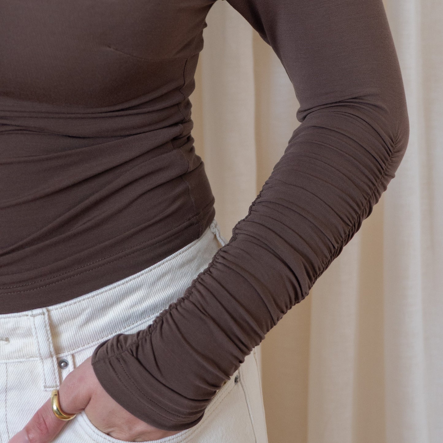 Brown long-sleeve top worn by a woman with a neutral background. Sinched sleeve detail with beautiful detailing. 